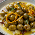 A rustic bowl of mixed marinated olives with orange peel, garlic, rosemary, and red pepper flakes on dark wood.
