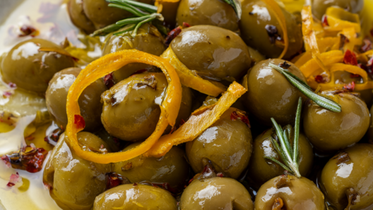 A rustic bowl of mixed marinated olives with orange peel, garlic, rosemary, and red pepper flakes on dark wood.