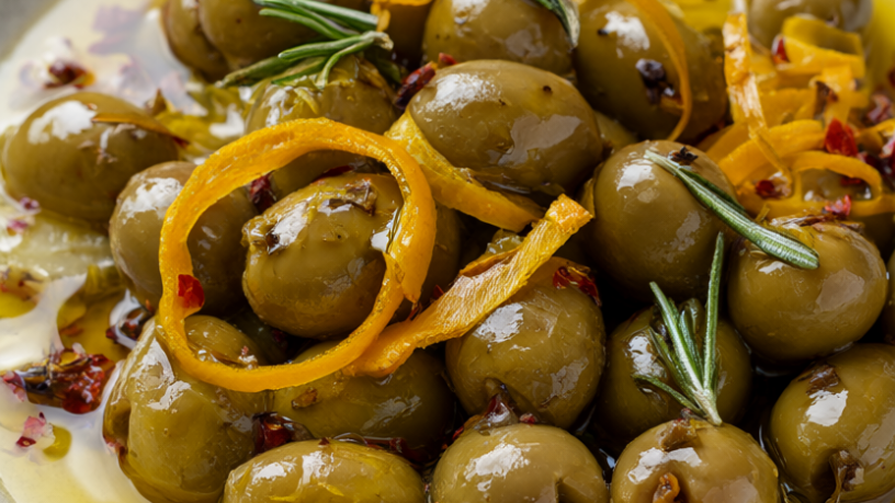 A rustic bowl of mixed marinated olives with orange peel, garlic, rosemary, and red pepper flakes on dark wood.