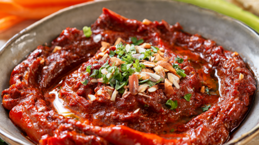 Roasted red pepper almond romesco dip in a ceramic bowl topped with olive oil, parsley and toasted almonds, with cucumber and carrot sticks and pita on a wood table