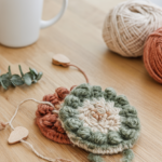 Two handmade punch needle coasters with woolly sage green, cream and terracotta loops on a pale oak table with a white mug, yarn and a wooden punch needle.