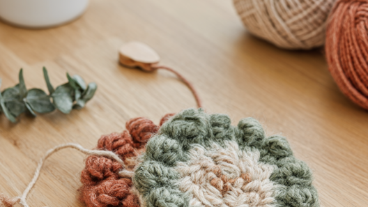 Two handmade punch needle coasters with woolly sage green, cream and terracotta loops on a pale oak table with a white mug, yarn and a wooden punch needle.