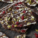 Broken pieces of dark chocolate bark topped with pepitas, cranberries, white chocolate drizzle and flaky salt on dark wood.